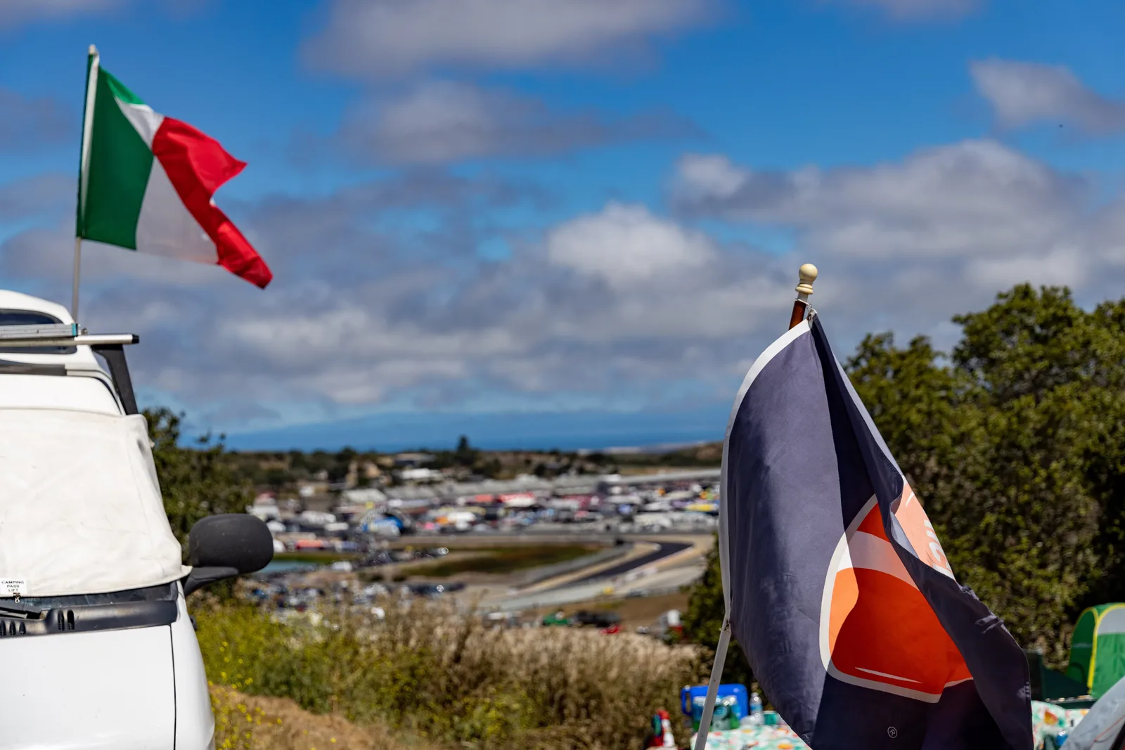An Italian flag and a navy and orange flag in the foreground, with the Laguna Seca circuit and paddock spread out in the valley below, July 2023