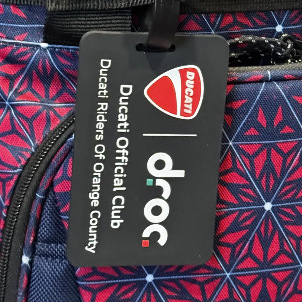 Black DROC luggage tag with the Ducati shield, the droc logo and the words Ducati Official Club, Ducati Riders Of Orange County, clipped to a backpack