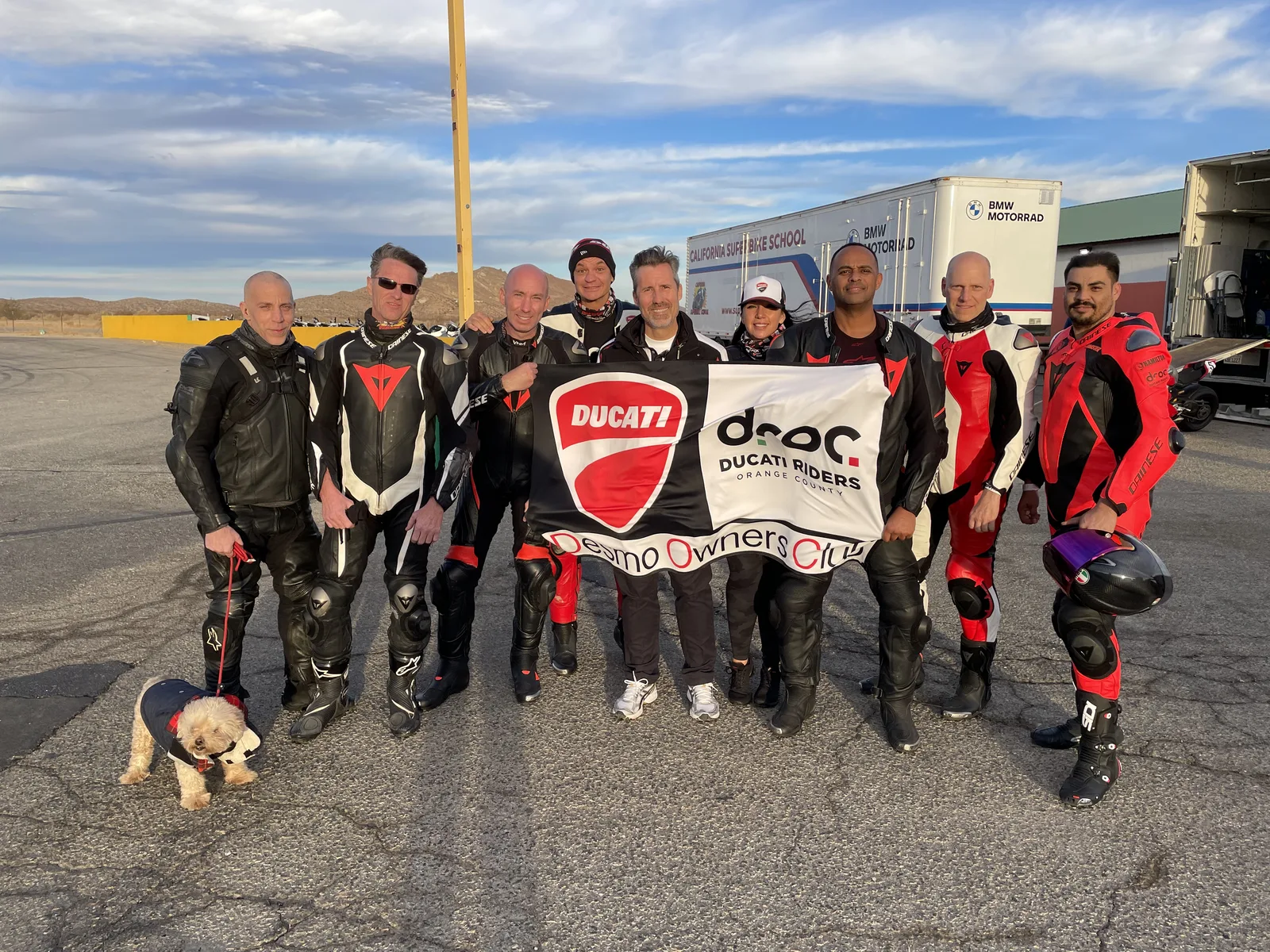 DROC track days at Streets of Willow 2 Eight Ducati Riders of Orange County members in leathers hold the DROC Desmo Owners Club banner on the pavement at Streets of Willow, with a small dog in front and the California Superbike School truck behind, October 2021