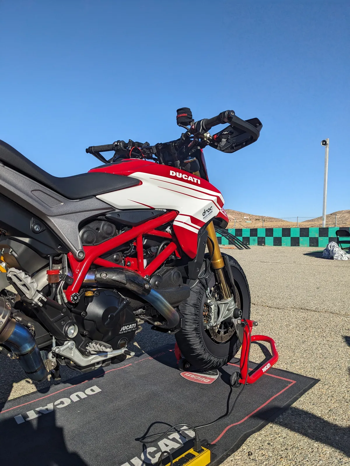 DROC track days at Streets of Willow 12 A red and white Ducati Hypermotard with DROC livery sits on a paddock stand with tire warmers on a Ducati mat at Streets of Willow, checkered wall and desert hills behind, November 2024