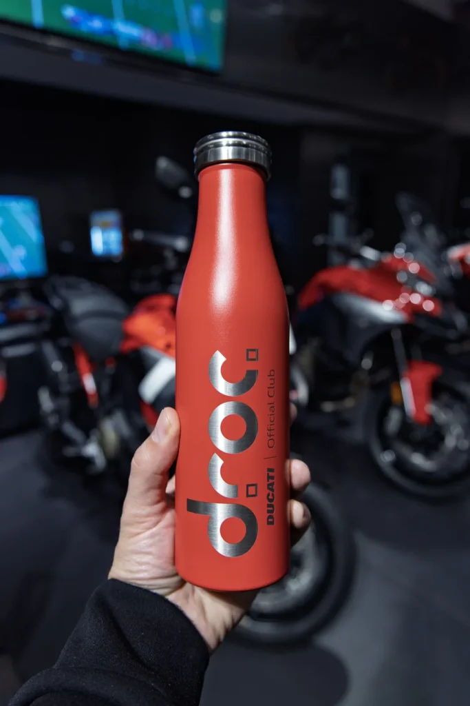 Red DROC Ducati Official Club water bottle held up in the Ducati Newport Beach showroom, motorcycles behind